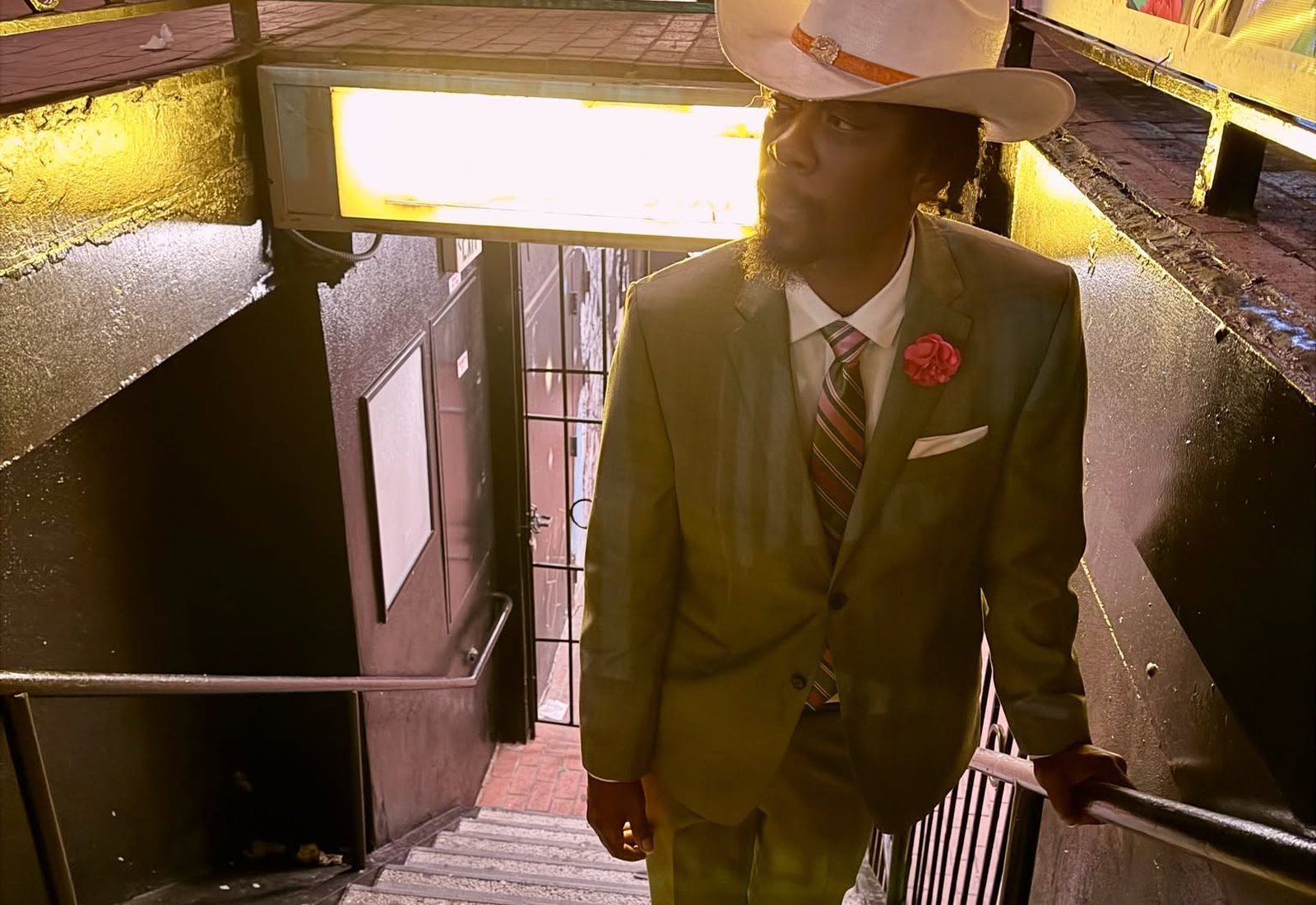 George Gentleman in an olive suit and a Panama hat, walking up a lit stairwell at night