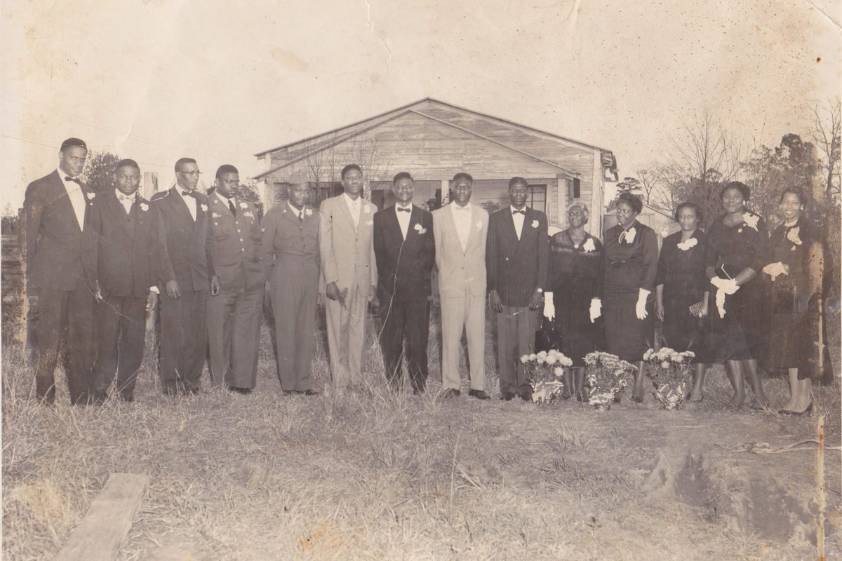 Vintage family portrait: a row of men in suits, bow ties and boutonnieres and women in dark dresses, white gloves and corsages, standing in front of a wooden house